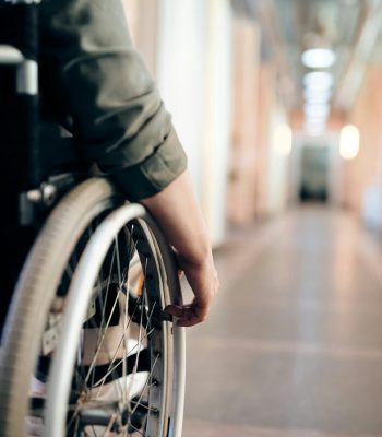 A person in a wheelchair is navigating an indoor hallway, focus on the wheel and hand.